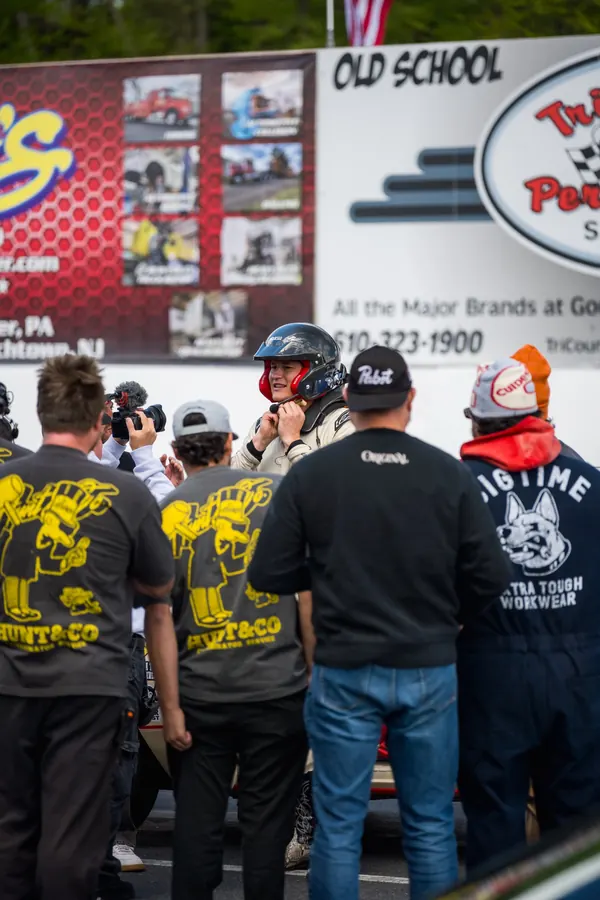 A driver in a helmet and race suit surrounded by crew and a camera operator at the starting line